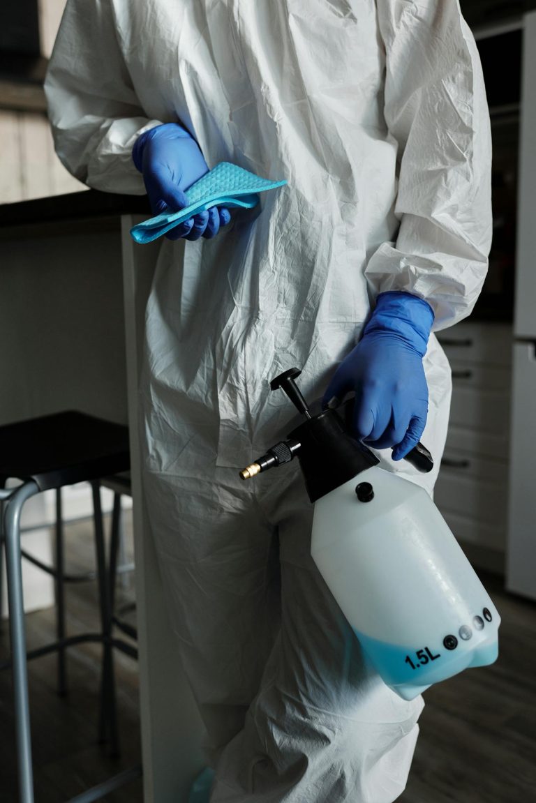 Individual in full protective gear holding disinfectant spray and gloves indoors for safety.