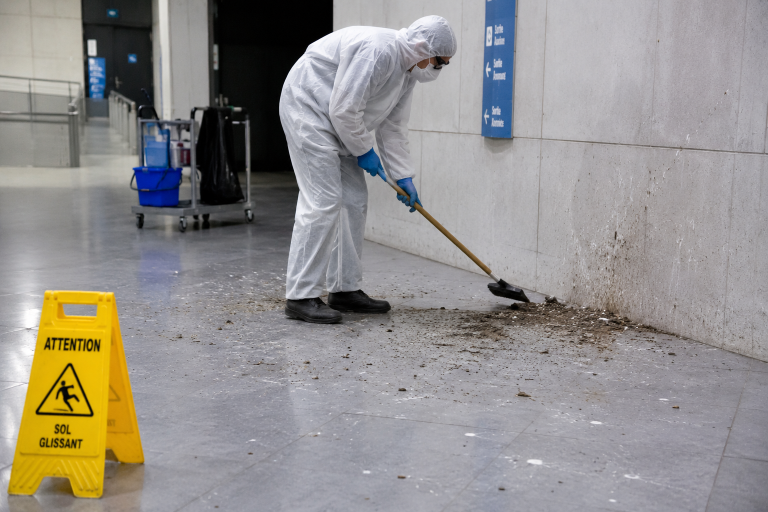 Agent équipé d’une combinaison de protection nettoyant des fientes de pigeon sur le sol d’un bâtiment public sécurisé par un panneau sol glissant.