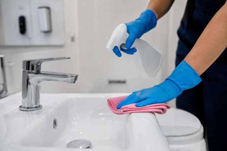 Personne avec des gants bleus désinfectant un lavabo blanc après le nettoyage d’une salle de bain