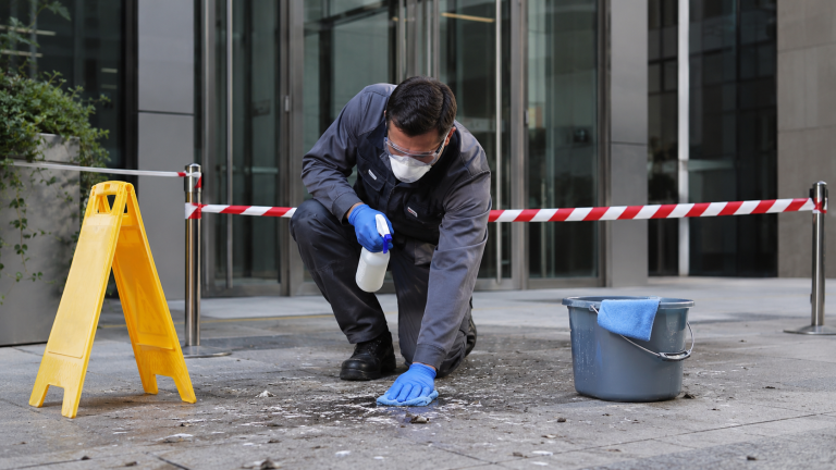 Technicien de nettoyage professionnel équipé de gants, masque et lunettes nettoyant des fientes de pigeon devant l’entrée d’un bâtiment d’entreprise sécurisé par un balisage.